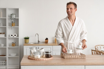 Young man in bathrobe putting cups of coffee on tray at home