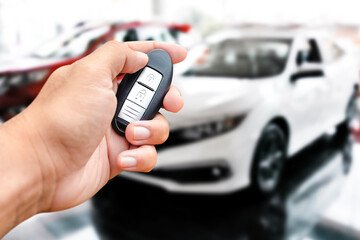 A hand holds a car key fob, with a white car blurred in the background. Car remote, vehicle access, keyless entry.
