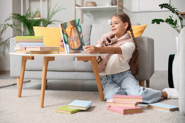 Cute schoolgirl reading book and sitting on carpet in living room