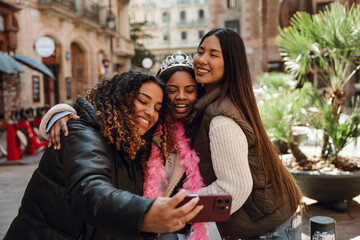 Three young women take a selfie outdoors on a city street. A Black woman wearing a crown and pink feather boa embraced by her Asian and Hispanic friends during a casual birthday celebration.