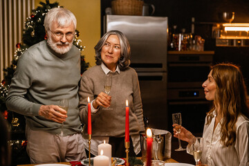 During a Christmas family dinner, grandparents stand beside the table with glasses of champagne while their daughter watches affectionately. A concept of family warmth, togetherness, and holiday.