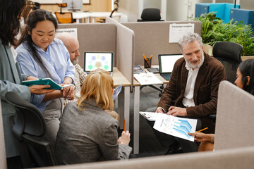 A senior White male office worker in a brown blazer is sitting in a shared office space, discussing printed charts with a multinational group of his colleagues, surrounded by laptops and documents.