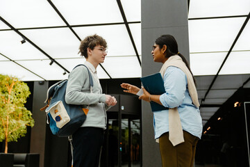 A young White man in a light gray hoodie and backpack listening to a young Indian woman in office attire holding a folder as they talk in a corporate building lobby during an internship discussion.