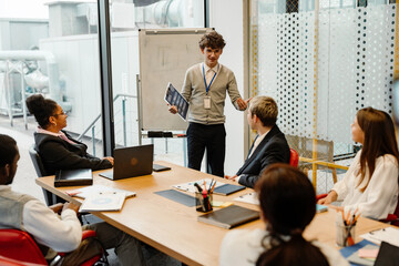 A young White man in a cardigan presenting printed materials to a multinational group of young people seated around a conference table during an internship meeting in a modern glass-walled office.