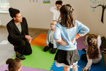A multiracial group of young children and a female teacher interact on colorful foam mats during a primary school classroom activity.