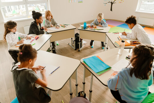 A multiracial group of young students sitting at desks in a modern classroom focused on drawing with colored pencils during an elementary school activity with their teacher.