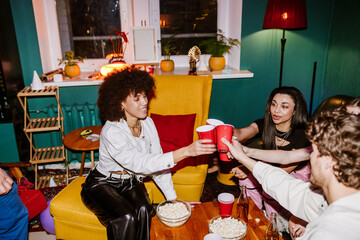 A multinational group of young people making a toast with red cups in a colorful, cozy living room...