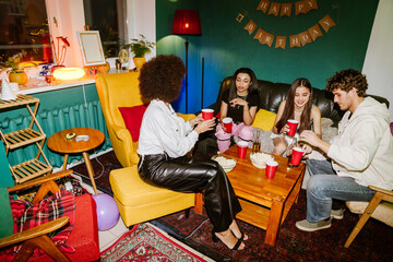 A multinational group of young people sitting in a cozy apartment living room decorated for a birthday party, sharing drinks and popcorn around a table with festive lighting and colorful furniture.