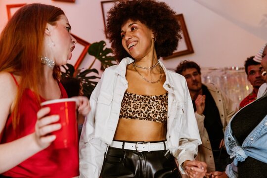 A multinational group of young people dancing and laughing during a vibrant birthday party in a decorated room featuring stylish outfits, red party cups, framed wall decor, and joyful expressions.
