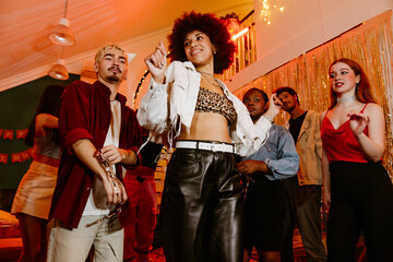 A multinational group of young people dressed in stylish outfits dancing and posing under warm party lighting in a decorated room with gold streamers and banners during a birthday celebration.