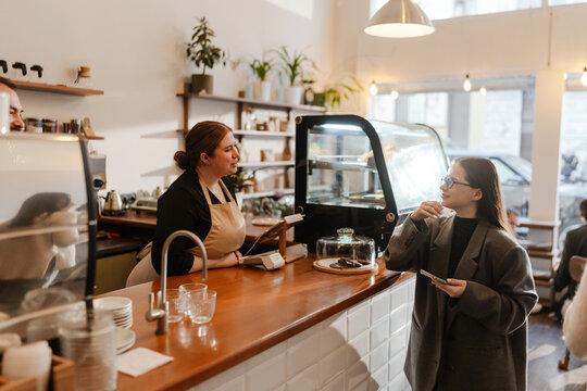 An adult woman wearing a coat and glasses talking to a barista behind the counter in a modern cafe, standing near the pastry display and holding a phone.
