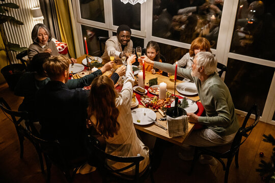 A group of Black and White people, including children, adults, and elderly people, gathers around a candlelit wooden dinner table. They smile and raise glasses in a festive toast.