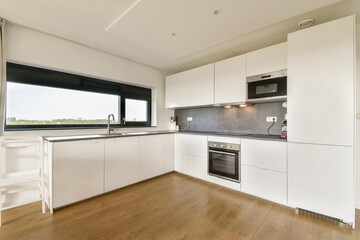 A sleek modern kitchen featuring white cabinetry, a grey countertop, and wooden flooring, with natural light streaming through a large window.