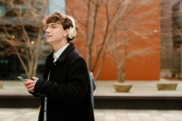 A young White man with glasses and headphones stands outdoors near a brick building, holding a...