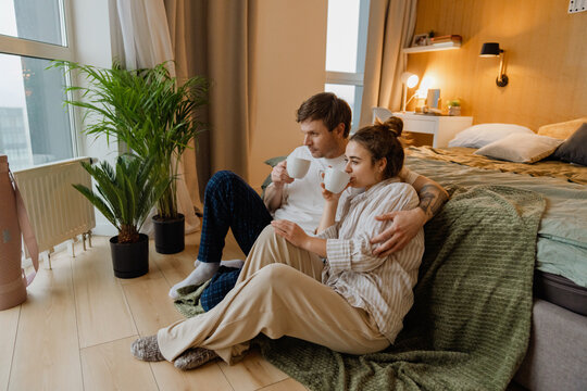 A young White couple shares a peaceful moment, sitting on their bedroom floor near a window. They lean against the bed, both sipping drinks from white mugs, enjoying quiet time together indoors.
