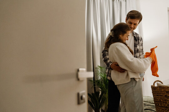 A view from the side of a tall White husband aged 30, hugging his pregnant White wife, who is in her late 20s, around the waist as they stand in a cozy bedroom, with her folding baby clothes - Powered by Adobe