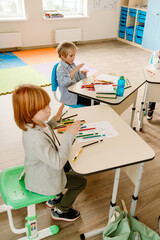 Obraz premium A school-aged Caucasian boy with long red hair, wearing a gray suit, sitting at his desk in a modern classroom and looking to the side, as the desks are arranged in a circle with his classmates
