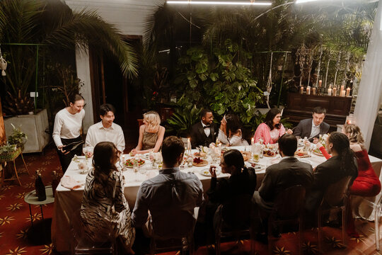 In a plant-filled wedding conservatory, a multiethnic group of guests sit talking at a long candlelit dinner table with a marrying Black couple in their 20s, while a White female server pours wine.