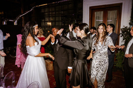 A young Black bride and groom dance and greet guests during their wedding reception in a dimly lit room. The diverse crowd of young and older people joins the celebration.