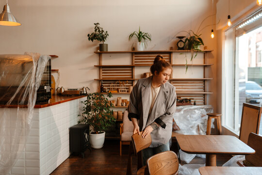 A young White female cafe owner wearing a grey blazer prepares a cafe space for opening. She carefully positions a wooden chair near a table while other furniture remains covered.