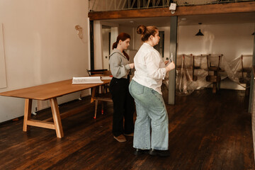 A view from the side of an overweight White waitress aged 25 and her thin White female colleague, walking around an empty coffee shop where they work and discussing renovations