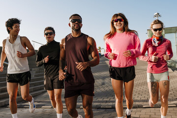 A handsome Black man in his early 30s in a brown T-shirt, two young European women in pink sweaters, an Asian adult man, and a young European man are running along a city street in sunny weather.