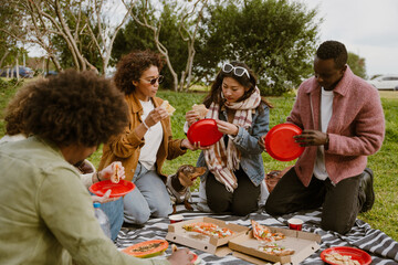 Diverse friends, Black, Latino, and Asian, enjoy a picnic sitting on a blanket in a park with grass. They share pizza slices and fruit from red plates, with their small dachshund joining the meal.