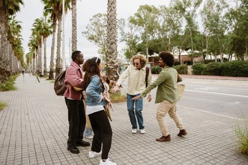 On a sidewalk lined with palm trees, five multiethnic friends in their 20s, two Black men, a White man, an Asian woman holding a dachshund dog, and a woman behind them, dance or play some game.