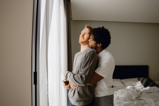 A view from the side of a loving gay couple in their 30s of a Black man and his White husband, hugging lovingly as the White man stands near the window at home and the Black man hugs him from behind - Powered by Adobe