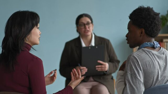 Rack focus of young biracial female psychologist trying to de-escalate high-conflict situation between emotional Asian teen girl arguing with Black boy during therapy consultation in class