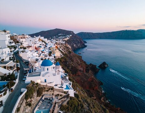 Low-altitude drone view of Mediterranean cliffside at magic hour, whitewashed villas with blue domes overlooking calm