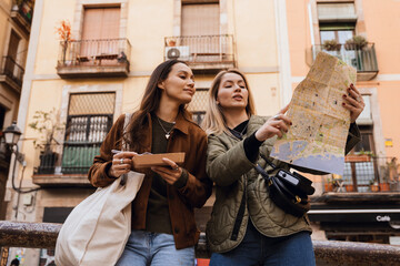 A Caucasian blond woman in her 20s, showing a paper map to her young Caucasian female friend as they're standing on a city street, with the friend eating some street sweets, on a sunny day