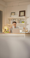 An elderly Asian man sits at the kitchen counter with a laptop and coffee mug, focusing on work or browsing during a calm morning at home. healthy retirement lifestyle, daily routine.