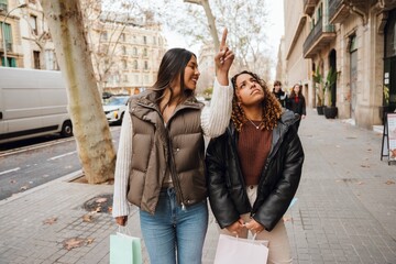 Two young friends, one Latino, and one Asian, walk along a city street with buildings and cars carrying shopping bags. One points up excitedly while the other looks, enjoying their day together. © Drobot Dean