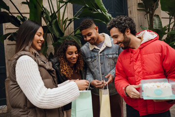 A multiethnic group of adult friends consisting of two women and two men holding paper bags and a cake while standing on the city street in front of an old building with green trees