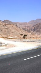 Road cuts through arid mountains, terrain fades into light beige textures, shadows of ridges deepen behind lane lines, rocks softly shaped by sun. Driving valley route.