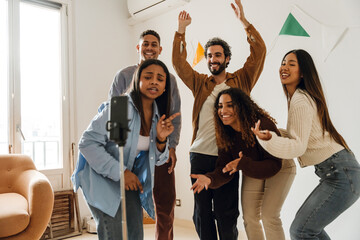Five diverse friends pose for a smartphone video recording indoors. They laugh and gesture, creating energetic content together at a house party in a room with white walls and a window.