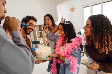 A group of young adult friends of different ethnicities standing around a Black woman in her 20s in a crown and a pink feather scarf with a birthday cake with candles in a room with a big window