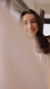 A cheerful woman with dark hair adjusts her hairstyle while looking at her reflection in a mirror, getting ready with confidence.