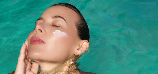  Relaxed woman floating in turquoise blue water with eyes closed, applying spf cream to her cheek....