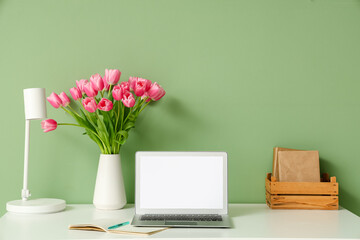 Stylish workspace with modern laptop, books and bouquet of beautiful tulip flowers near green wall