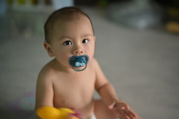 A curious baby with a pacifier sits on the floor, holding a toy, surrounded by a softly blurred...