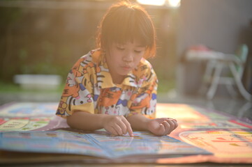 A young child in a colorful shirt is engrossed in a book, sitting on a mat in a sunlit outdoor...