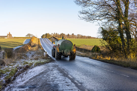 Tractor with a Slurry Tank Driving on a Wet Country Road with Frost on Ground in Shadow