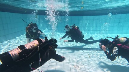 Scuba divers engaged in underwater training session actively in swimming pool. Divers in process of adjusting goggles as colleagues continue with gesticulating practice