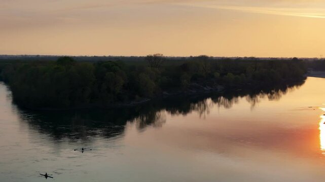drone pans gently over the Po River near Olza, capturing rowers moving through calm golden waters beside dense tree lines, mirrored reflections and subtle motion evoke poetic stillness, slow motion.
