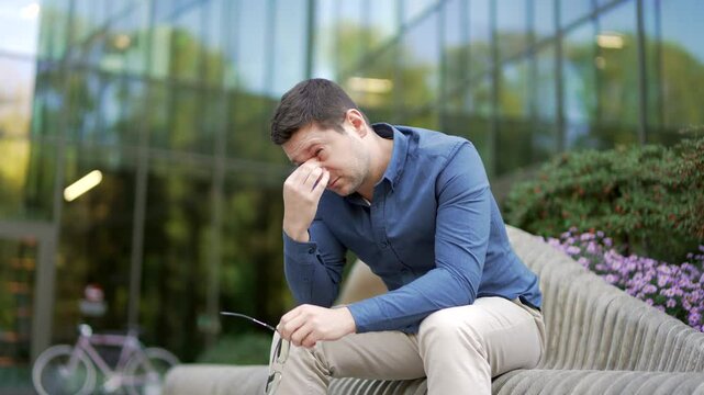 Businessman suffering from eye strain while sitting on bench on street near modern office building. Upset adult handsome man worker in a blue shirt takes off glasses and massages, rubs bridge of nose