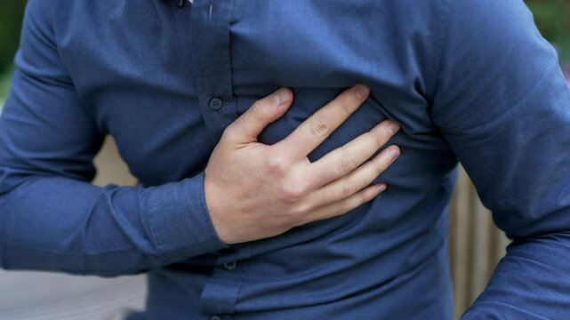 Close up. Male hands holding chest. Man in blue shirt has heart attack sitting on bench on street near business office building. Sick man massaging a painful sore spot, needs urgent medical attention