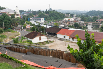 Obraz premium Cracked asphalt road showing damage from potential landslide or subsidence. Temporary retaining wall in place, overlooking a town and river.