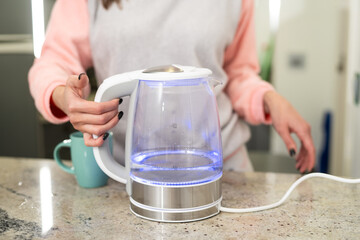 Woman in kitchen holding boiling modern glass electric kettle.